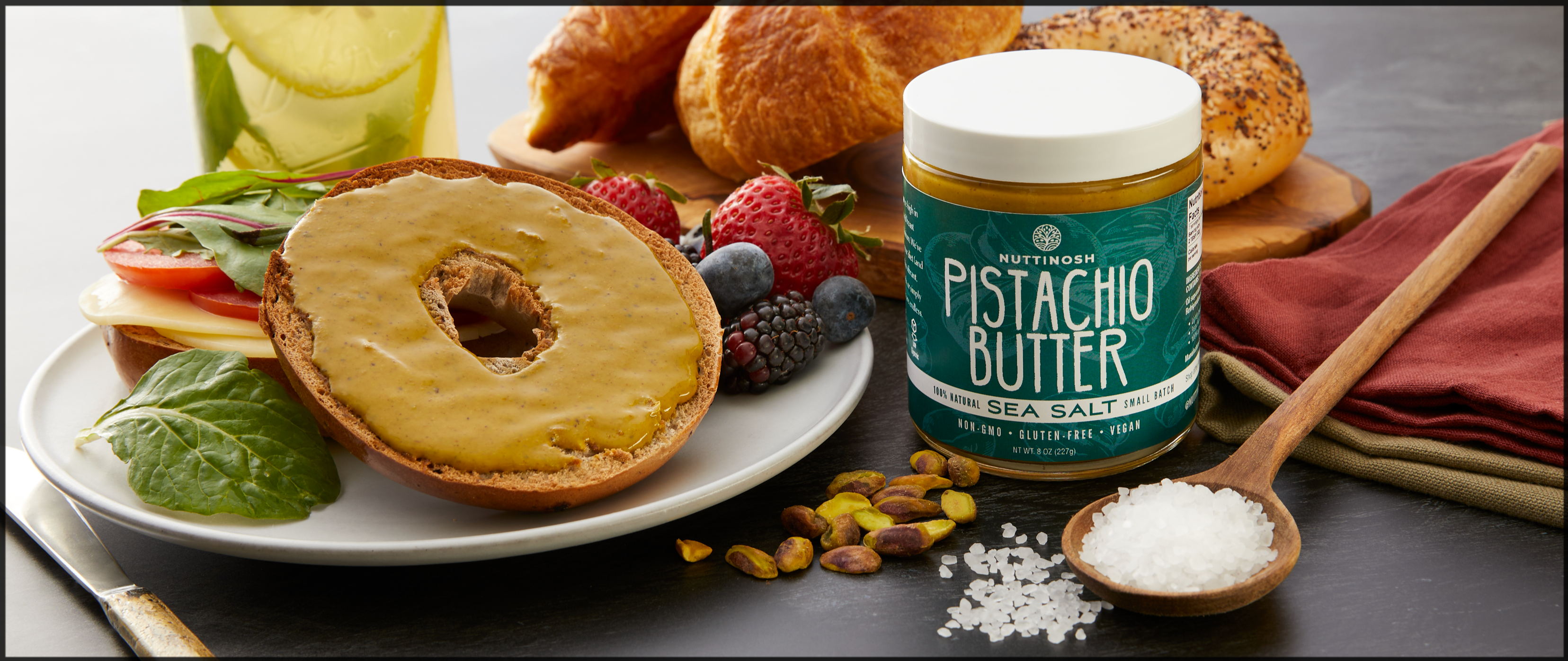 An assortment of fresh fruit, breakfast bagels, and breads with a jar of Nuttinosh pistachio butter in the center.