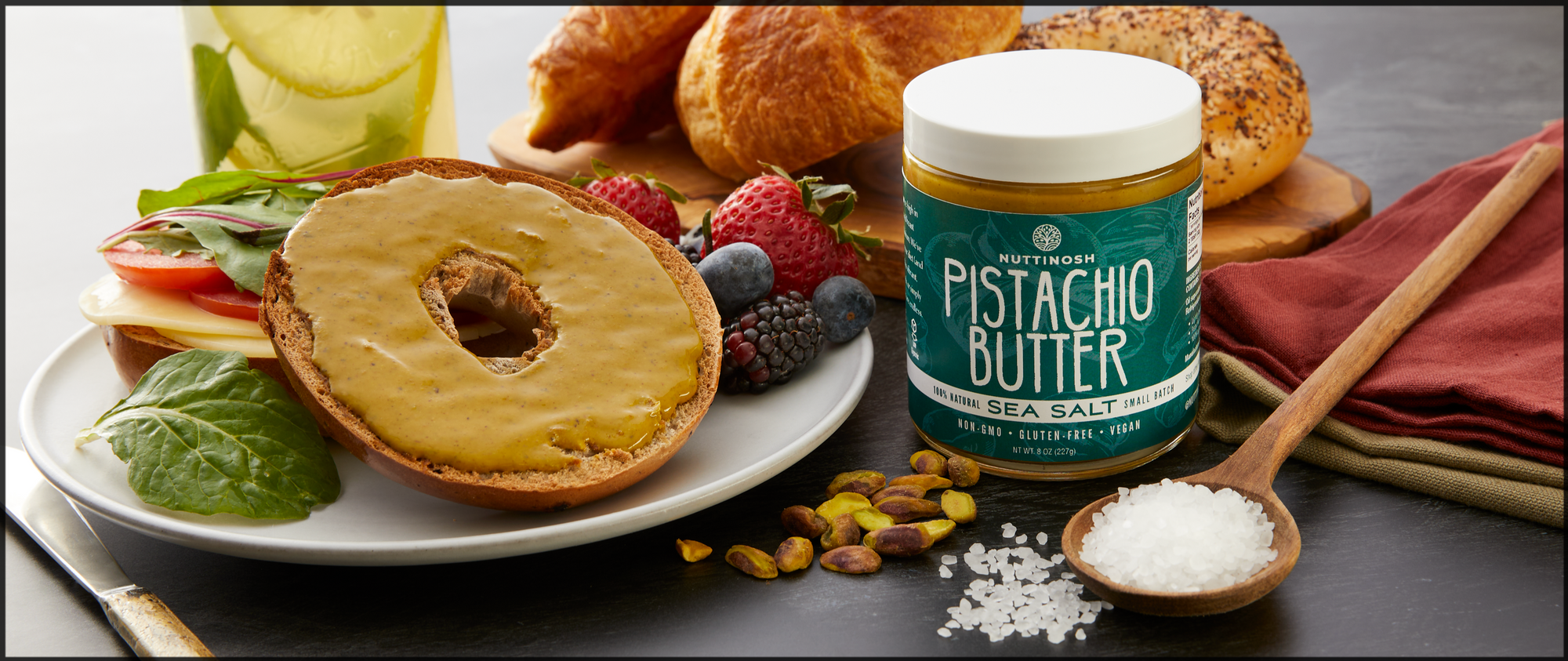 An assortment of fresh fruit, breakfast bagels, and breads with a jar of Nuttinosh pistachio butter in the center.