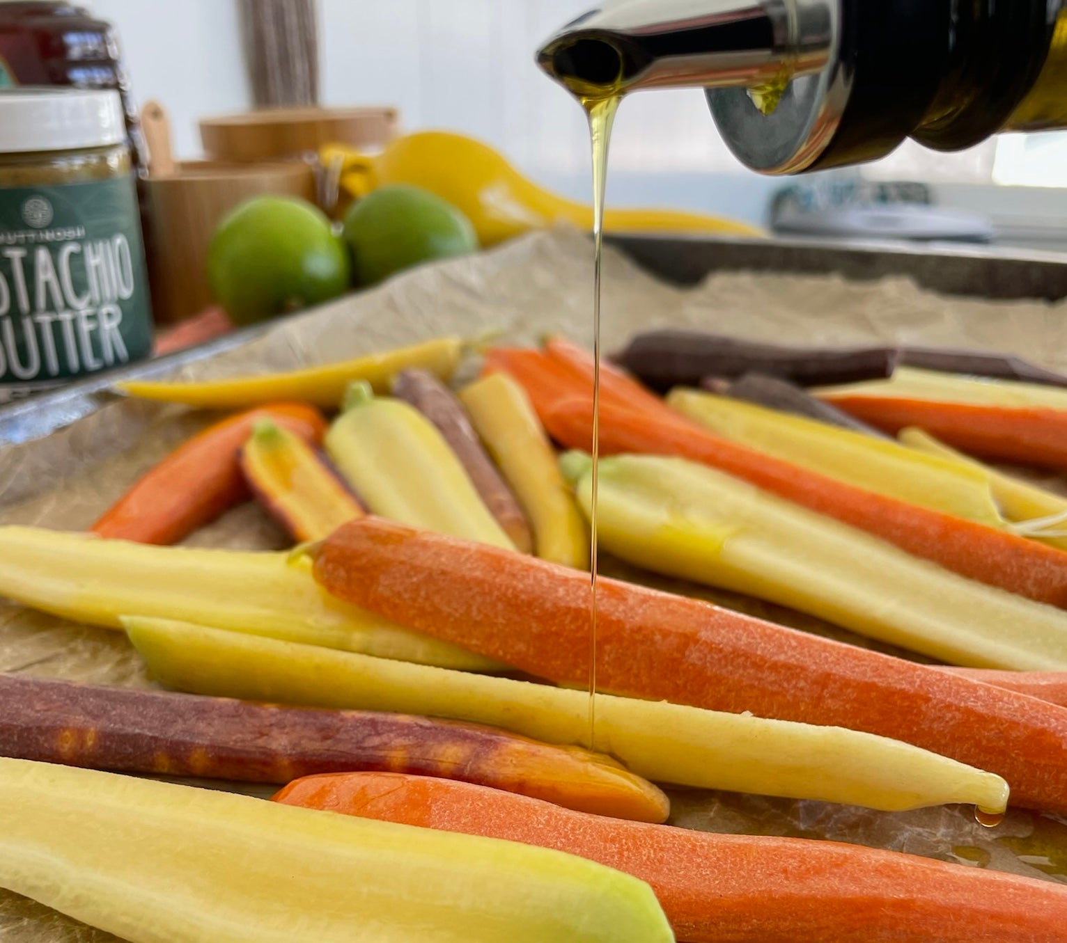 A baking sheet lined with fresh cut multicolored orange, yellow, and purple carrots. Olive oil and pistachio butter is being drizzled over the carrots.