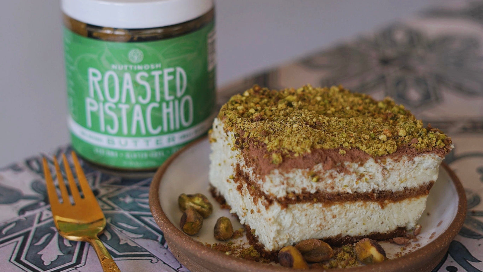 Slice of pistachio tiramisu topped with crushed pistachios beside a jar of Nuttinosh roasted pistachio butter