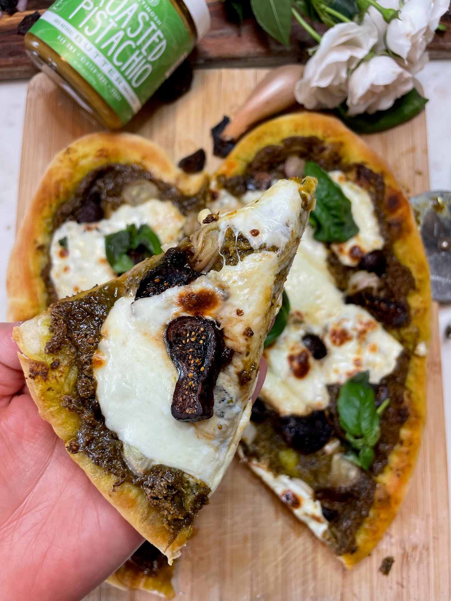 A freshly baked heart shaped pistachio butter pesto pizza with figs, mozzarella and balsamic glaze as toppings. A hand is holding a slice of the pistachio butter pesto pizza.