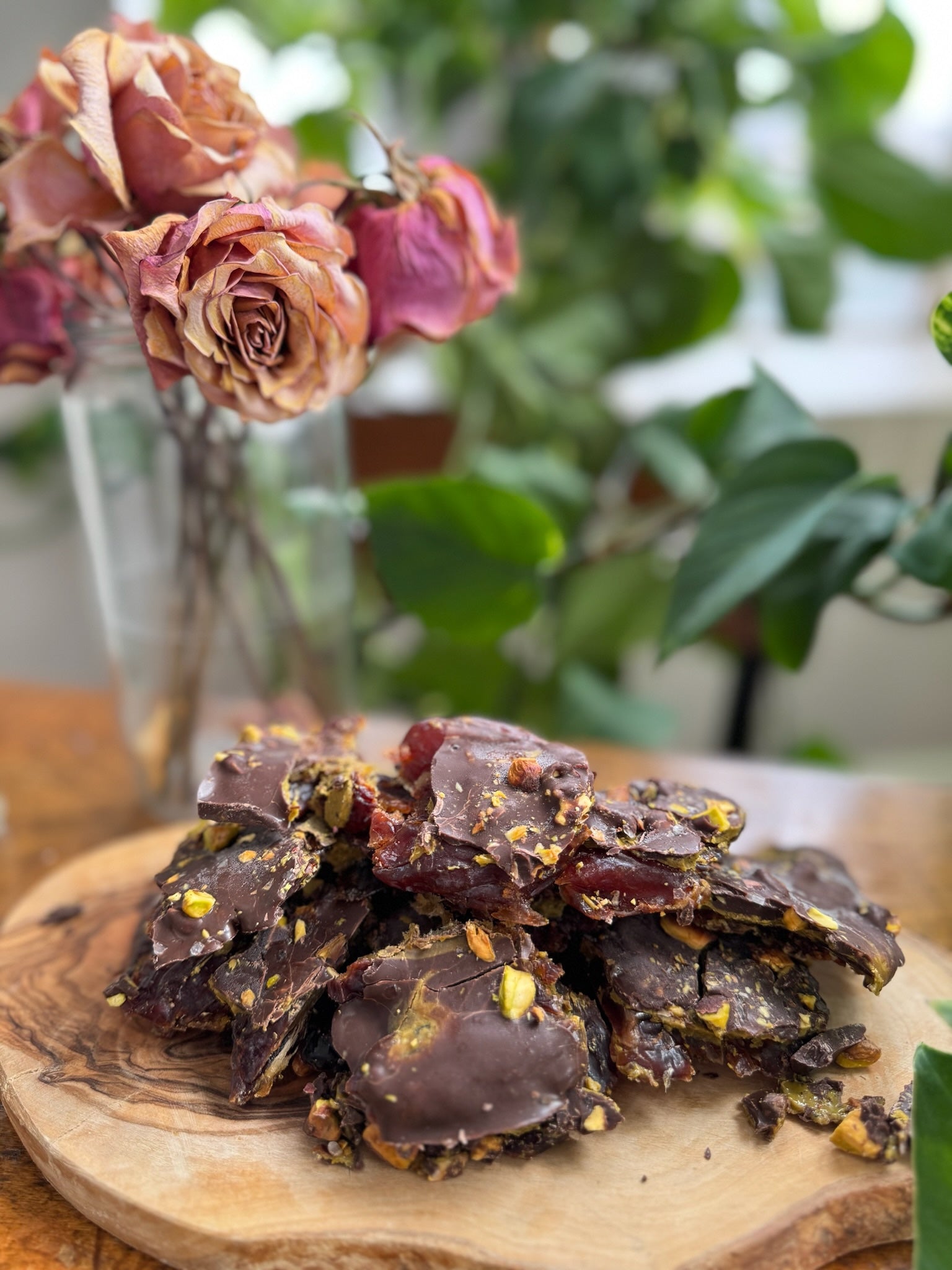 No-Bake Pistachio Date Bark stacked on a wooden plate with bright pink dried flowers and green leaves in the background.