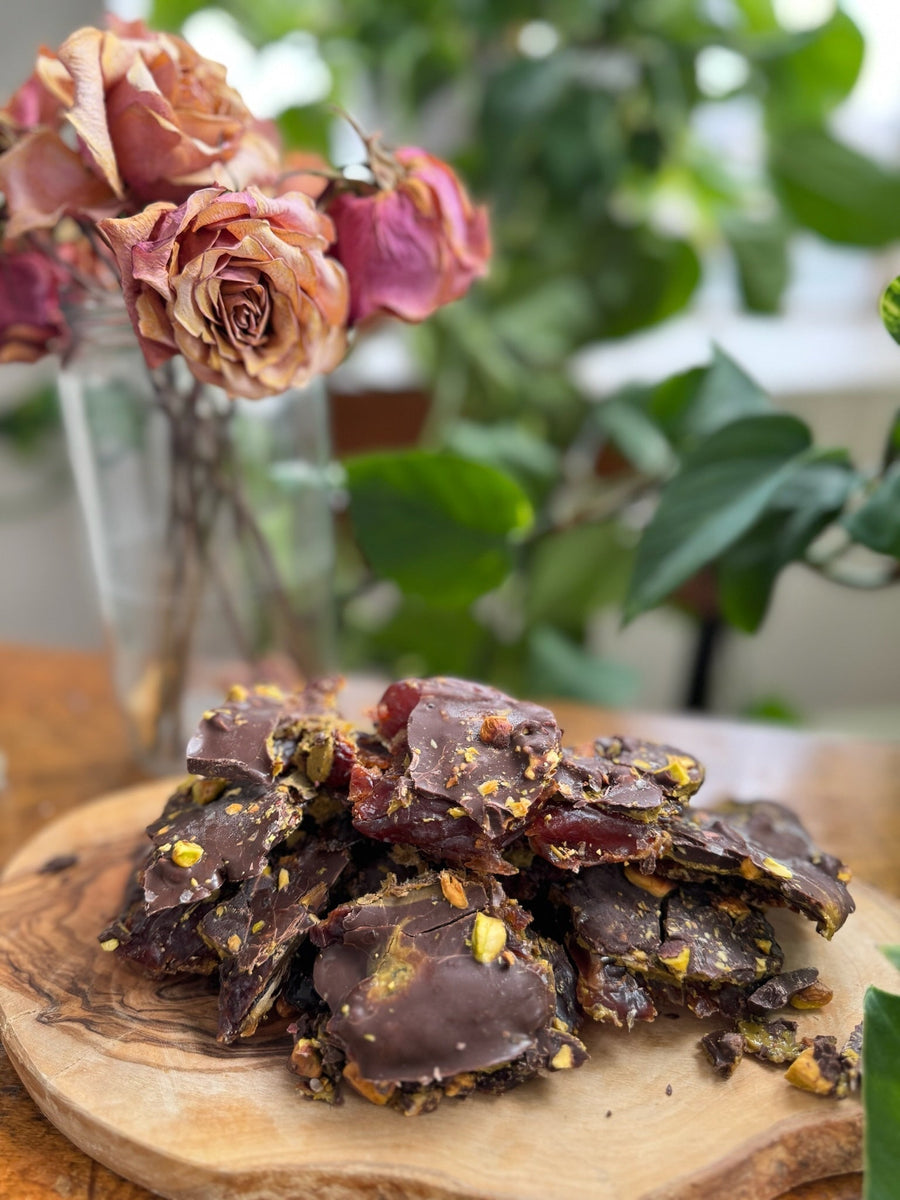 No-Bake Pistachio Date Bark stacked on a wooden plate with bright pink dried flowers and green leaves in the background