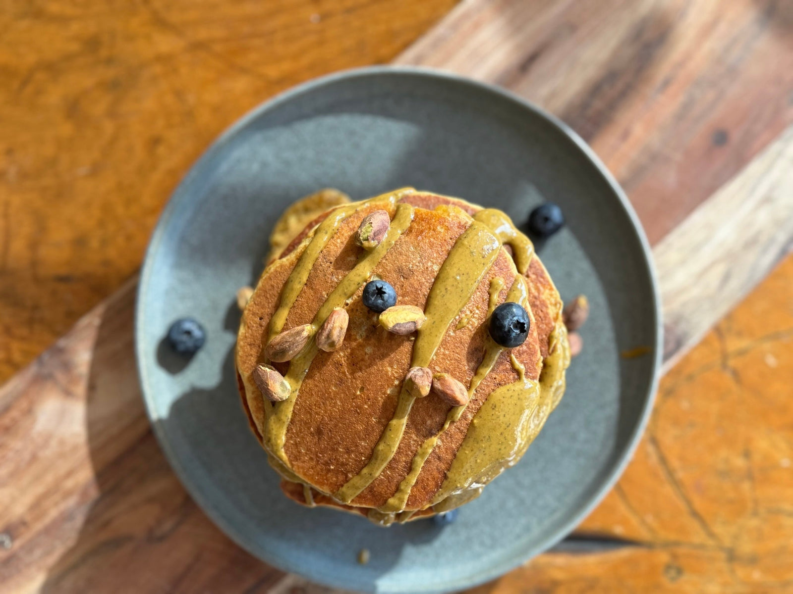 A stack of fluffy pistachio butter oat pancakes on a blue ceramic plate. The yare topped with fresh blueberries, pistachio butter, and whole unshelled pistachios