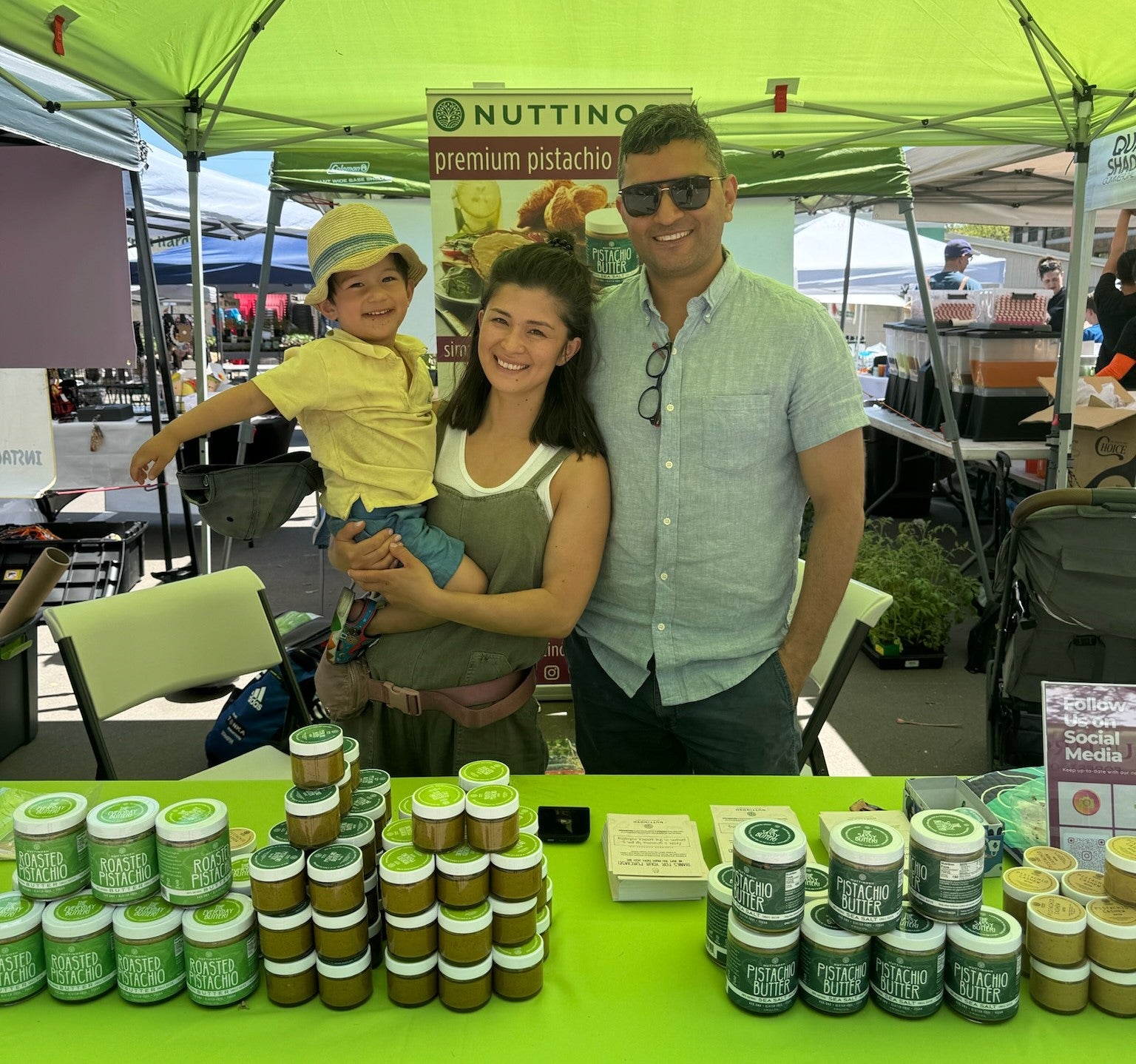 Nuttinosh founder Anosha Singh and her family standing behind a table full of stacked Nuttinosh pistachio butter jars at the farmer's market in Minneapolis, Minnesota.