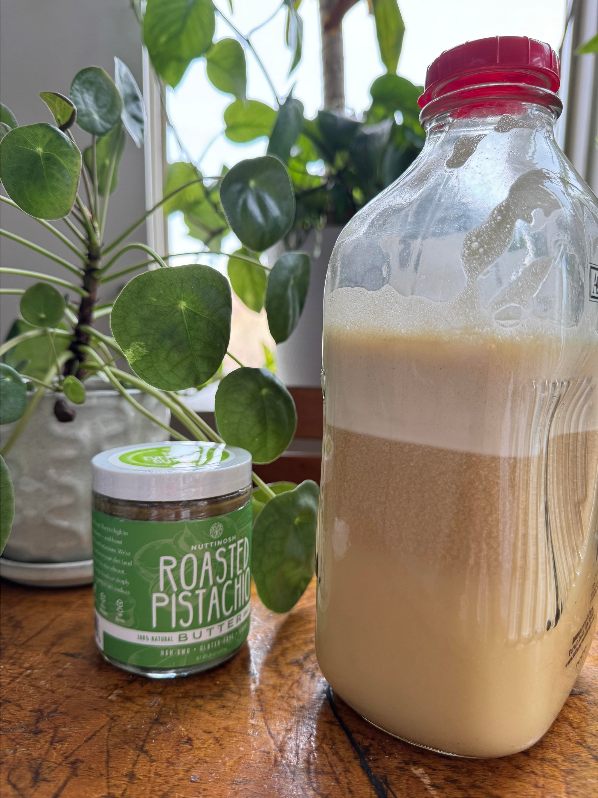 A glass milk jug of freshly-made pistachio nut milk stands on a wooden countertop. A jar of Nuttinosh Roasted Pistachio Butter sits on the countertop next to the pistachio milk.