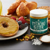 Jar of Nuttinosh Pistachio Butter Sea Salt next to a bagel spread with pistachio butter, fresh berries, croissants, and a spoonful of sea salt on a breakfast table.